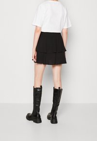 Osoba oblečená v bílém krátkém rukávu, černé vrstvené mini sukni a černých vysokých botách, stojící na hladkém bílém pozadí.