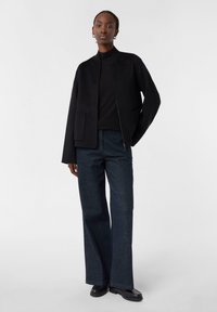 Schwarze Zip-Jacke mit hohem Kragen, großen Taschen, kombiniert mit dunkelblauen weiten Jeans und schwarzen Stiefeln. Glatte Textur.