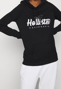 Person som bär en svart Hollister California hoodie med framficka och vita byxor, står med händerna på sina höfter.