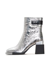 Metallic silver ankle boot with a textured surface, block heel, and a black accent strap featuring a logo detail. Shiny and smooth finish.