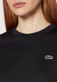 Gros plan d'une personne portant un t-shirt noir à col rond avec un petit logo crocodile vert et blanc sur la poitrine.