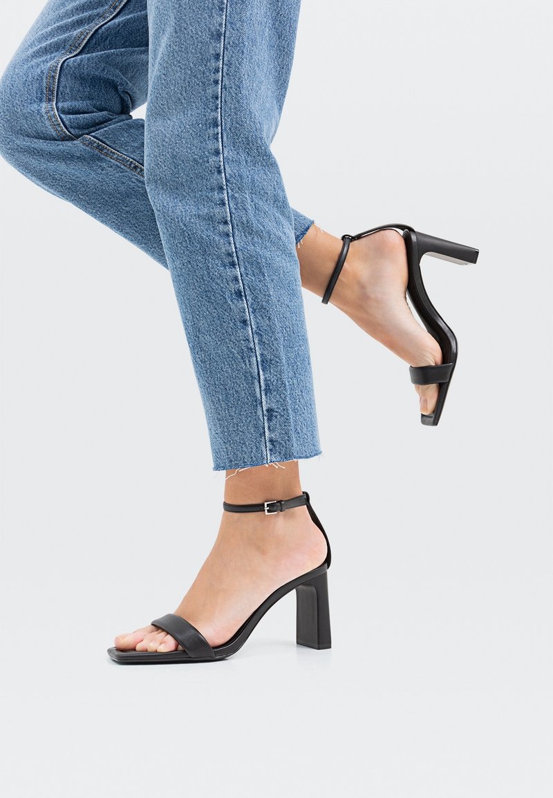 Femme portant un jean bleu et des sandales noires à talons hauts avec brides à la cheville, un pied à plat et l’autre levé sur un fond uni.