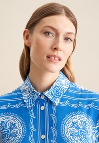Camicetta blu con un motivo paisley bianco, colletto button-down e tre bottoni. Tessuto liscio, maniche corte.