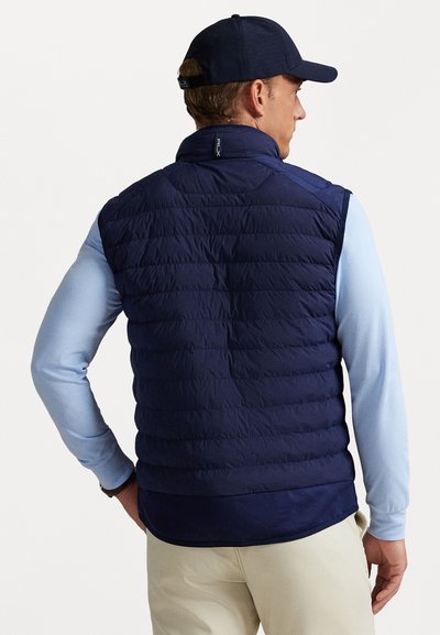 Man wearing navy blue quilted vest, light blue long-sleeve shirt, beige pants, and navy blue cap, facing away against a white background.