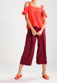 Červená off-the-shoulder blůza s volánkovými rukávy, kombinovaná s burgundskými širokými kalhotami a oranžovými sandály na podpatku.