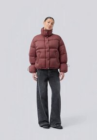 Frau trägt eine burgunderrote Pufferjacke mit hohem Kragen, schwarze weite Jeans und schwarze Schuhe und steht vor einem schlichten hellen Hintergrund.