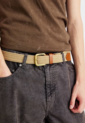 Ceinture enroulée en tan avec une boucle en or et un accent en cuir marron, portée avec un pantalon en velours côtelé gris et une chemise marron foncé.
