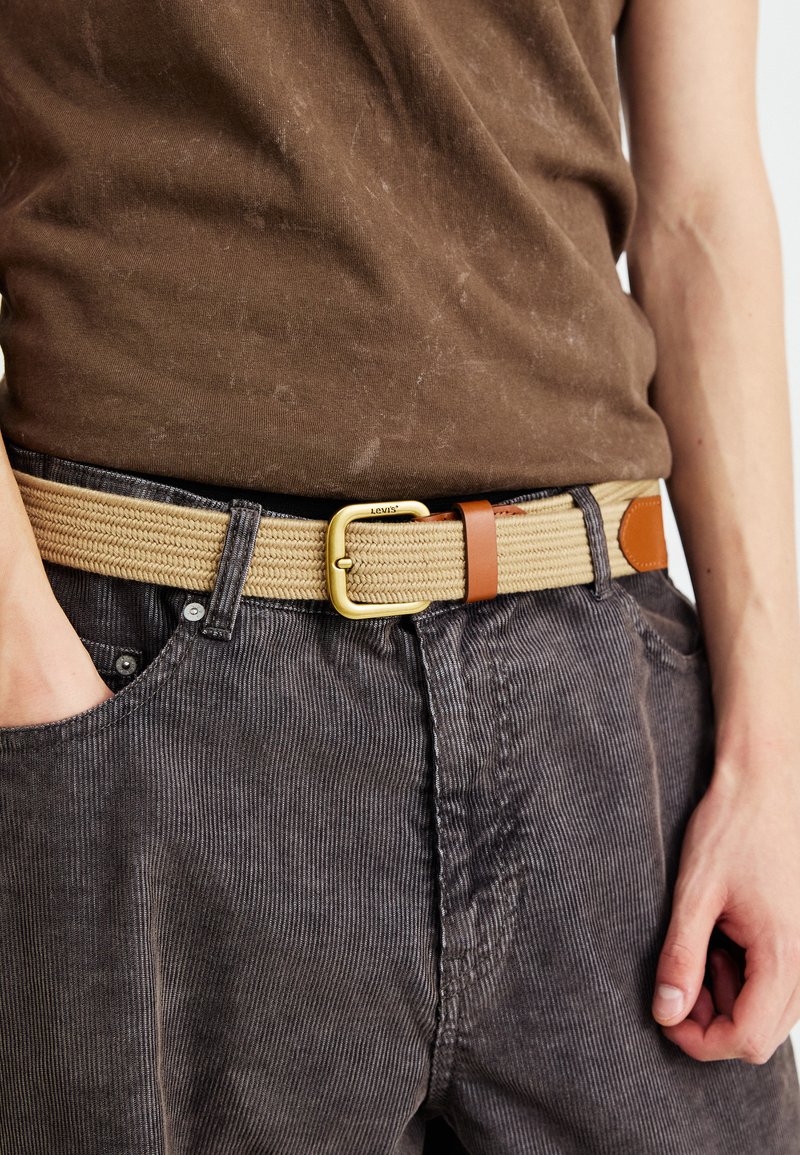 Cinturón de tejido color marrón con hebilla dorada y detalle de cuero marrón, usado con pantalones de pana grises y una camisa marrón oscura.