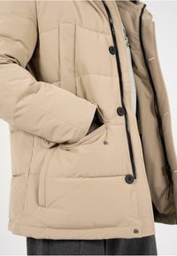 Giubbotto imbottito beige con chiusura a zip e bottoni neri. Comprende tasche laterali e un tessuto testurizzato per calore e comfort.