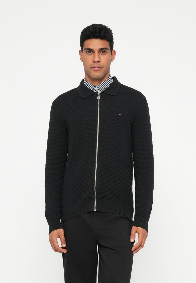 Fekete kötött, zippzáros kardigán gallérral, texturált anyaggal, bordás mandzsettákkal és derékkal. Fehér logóhímzés a mellrészen, kockás ing fölött.