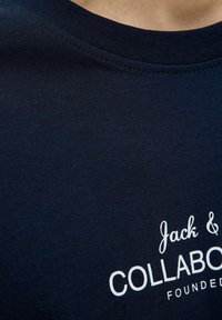 T-paita tummansinistä puuvillaa, jossa on pyöreä pääntie ja valkoista painatusta tekstiä, mukaan lukien "Jack &" ja "COLLABORATION", sekä sileä, teksturoitu pinta.