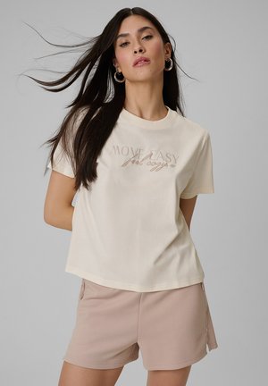 Kobieta o długich ciemnych włosach, ubrana w beżowy T-shirt z napisem „MOVE EASY feel cozy” oraz jasnoróżowe szorty na jednolitym szarym tle.