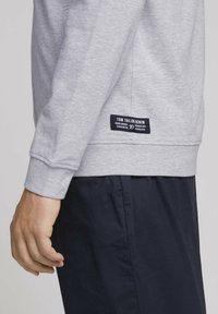 Grey sweatshirt made of cotton blend, featuring ribbed cuffs and hem, with a navy label on the lower side displaying “TOM TAILOR DENIM.”