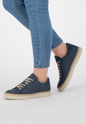 Navy blue suede sneakers with beige laces, round toe, and textured sole featuring a woven edge; styled with cuffed denim jeans.