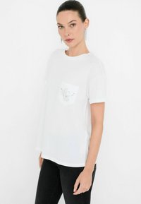 T-shirt bianco a maniche corte con tasca e dettagli decorativi argentati. Tessuto morbido con vestibilità rilassata, abbinato a jeans neri.