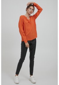 Helle orange Bluse mit langen Ärmeln, V-Ausschnitt und gerafften Schultern, kombiniert mit engen schwarzen Lederhosen und weißen Sneakers.