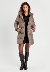 Pufferjas in taupe met capuchon, ritssluiting aan de voorkant en zijzakken; heeft een gestikte ontwerp en zachte textuur. Gekozen boven een zwart outfit.