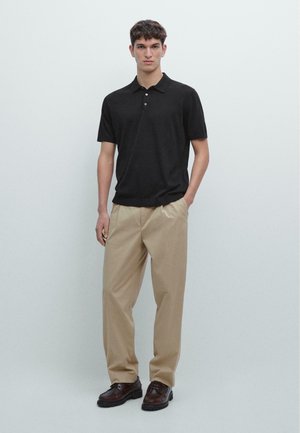 Junger Mann steht, trägt ein schwarzes Polohemd mit kurzen Ärmeln, beigefarbene Hose und dunkelbraune Lederschuhe, mit einer Hand in der Tasche.