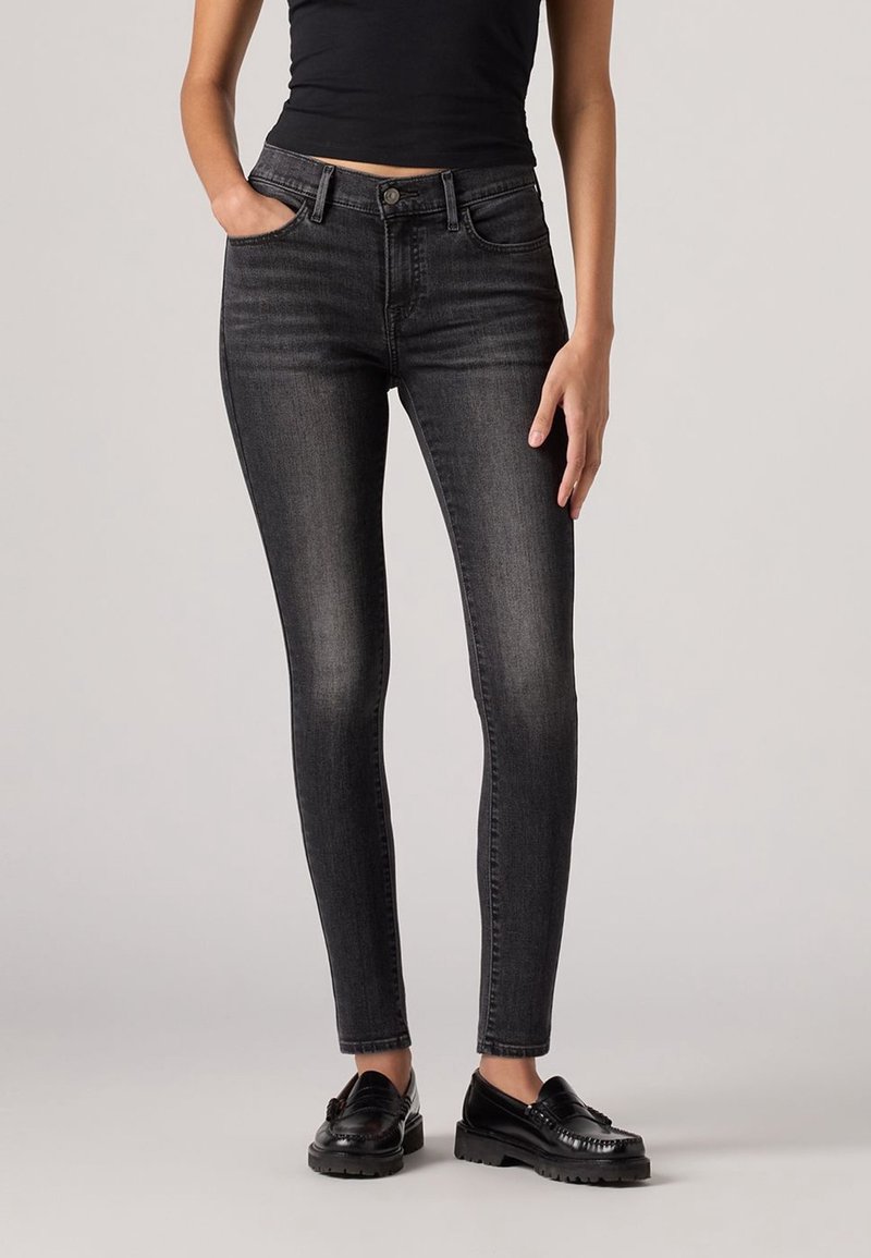 Femme portant un jean slim noir, un top sans manches noir et des mocassins noirs, debout avec une main dans la poche devant un fond uni.