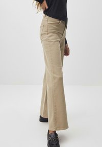 Beige Schlagjeans mit mittelhohem Bund, vorderen Taschen und kontrastierender Naht. Kombiniert mit schwarzen Slippern, seitlich dargestellt.