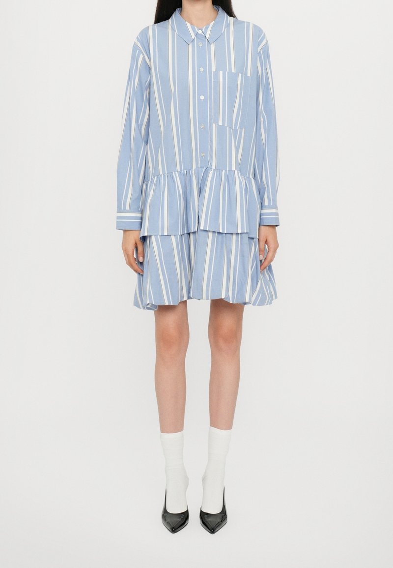 Femme portant une robe chemise à manches longues à rayures verticales bleu et blanc avec une poche avant, des chaussettes blanches et des talons pointus noirs.