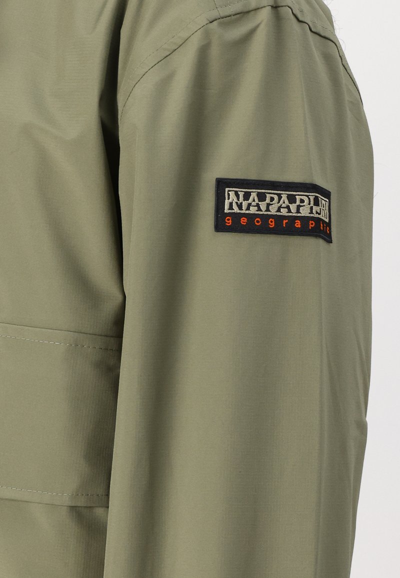 Manga de chaqueta verde oliva con un parche rectangular negro cosido que muestra el logo "NAPAPIJRI geographic" en texto blanco y naranja.