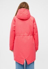 Persoon met lang haar in een felroze gewatteerde jas en blauwe jeans, gezien van achteren tegen een effen achtergrond.