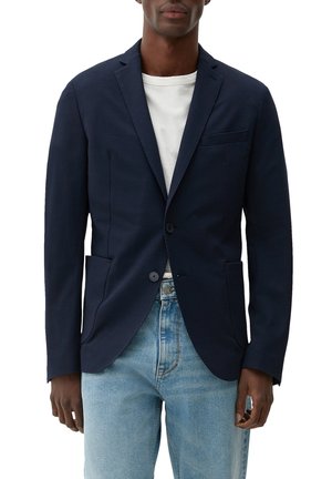Man draagt een marineblauwe blazer over een wit t-shirt met lichtblauwe spijkerbroek, staand tegen een witte achtergrond.