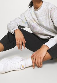 Mikina Nike světle šedé barvy s barevným mramorovým vzorem, kombinovaná s černými kalhotami a bílými teniskami. Měkká látka s žebrovanými manžetami.
