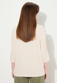 Hellbeige, lockere Bluse mit Dreiviertelärmeln und abgerundetem Saum, aus weichem, strukturiertem Stoff, von hinten präsentiert.