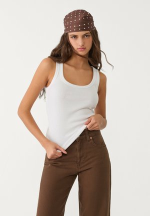 White ribbed tank top paired with brown wide-leg pants; model wearing a brown polka-dot headscarf. Neutral background.