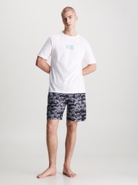 T-shirt bianca in cotone con logo CK azzurro chiaro, abbinata a pantaloni da bagno neri con un motivo astratto blu e bianco.