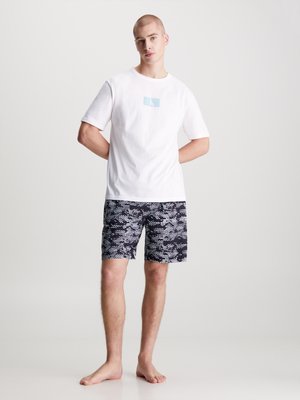 T-shirt bianca in cotone con logo CK azzurro chiaro, abbinata a pantaloni da bagno neri con un motivo astratto blu e bianco.