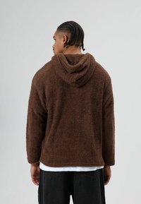 Veste polaire à capuche marron avec une surface texturée et duveteuse et des manches longues. Caractérisée par une coupe décontractée et une poche kangourou à l'avant.