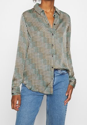 Mujer que lleva una camisa de botón de manga larga con un patrón de pata de gallo multicolor, metida parcialmente en unos vaqueros azules.