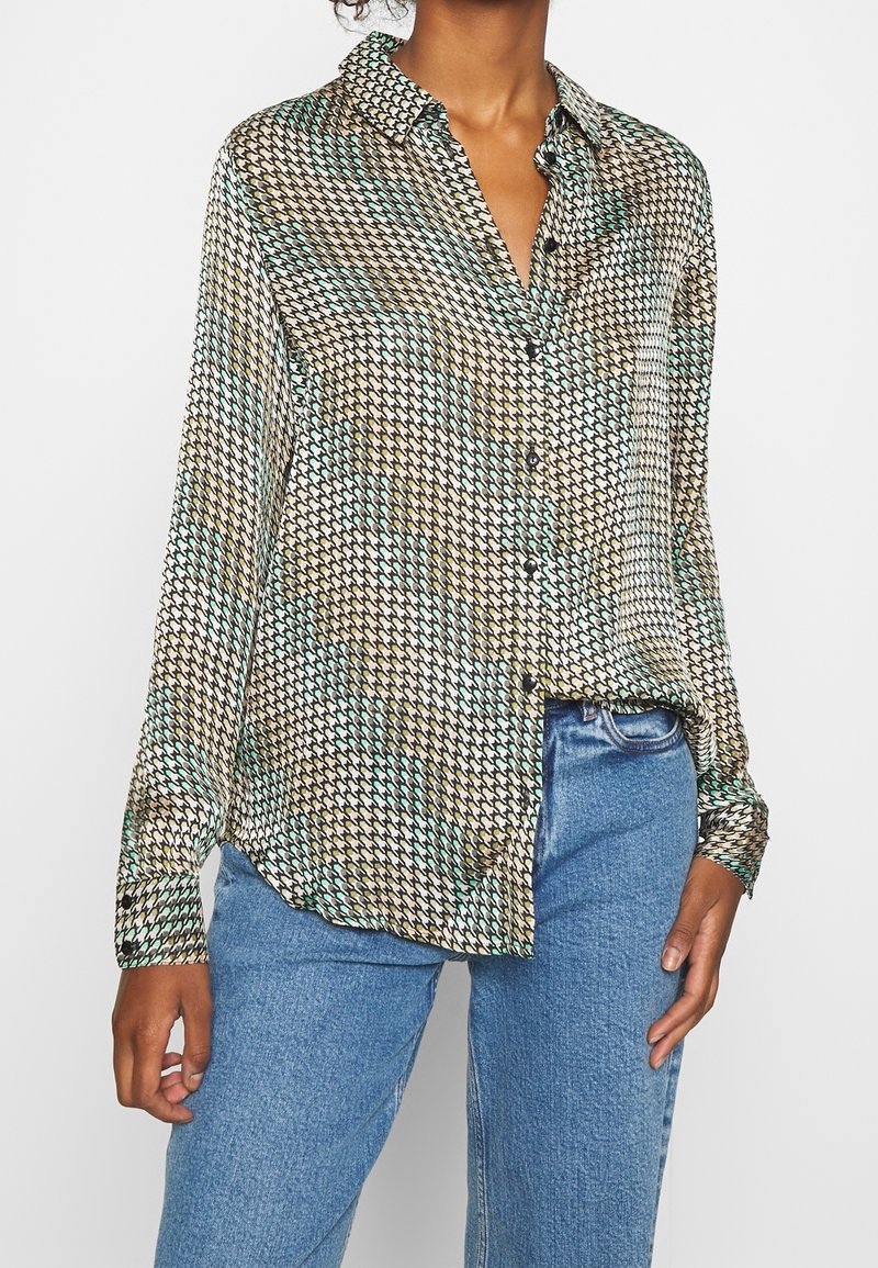 Woman wearing multicolored houndstooth patterned button-up shirt with long sleeves tucked partially into blue jeans.
