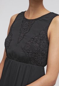 Svart klänning med en spetsbyxlinje med intrikata mönster, rund halsringning och en flödande kjol. Tyget verkar slätt och lätt.