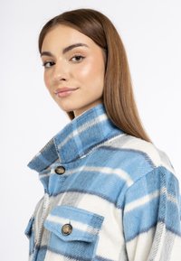 Blauwe en witte geruite fleecejas met een kraag, twee voorkolletten en knopenaccenten. De stof heeft een zachte, gestructureerde afwerking.