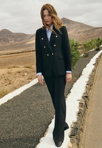 Czarna marynarka dwurzędowa z złotymi guzikami, zestawiona z czarnymi spodniami o dopasowanym kroju i jasnoniebieską koszulą. Gładki materiał; dopasowany fason.