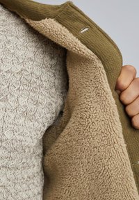 Ręka trzyma otwartą oliwkowo-zieloną kurtkę, która odsłania miękką, beżową podszewkę z polarowego materiału oraz teksturowany jasnoszary sweter dzianinowy znajdujący się pod nią.