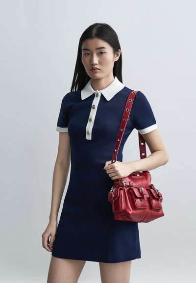 Jeune femme portant une robe bleu marine avec un col blanc et des boutons, tenant un sac à main rouge avec des poches avant et des détails de boucle.