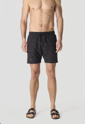 Hombre sin camiseta vestido con pantalones cortos de baño negros con sutiles patrones náuticos y sandalias negras de punta abierta, de pie frente a un fondo gris liso.