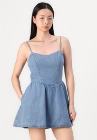 Denim obleka z tankimi naramnicami, ki ima oprijet zgornji del in široko krilo, v svetlo modri barvi z rahlo teksturo in stranskimi žepi.