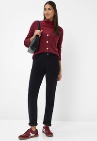 Burgunderfarbene Strickjacke mit Knöpfen, über einem kastanienbraunen Rollkragenpullover getragen, kombiniert mit schwarzen Cordhosen und burgunderfarbenen Turnschuhen. Schwarze Tasche.