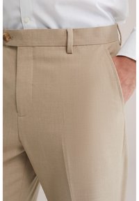 Nahaufnahme von beigen Anzughosen mit Seitentaschen, getragen mit einem weißen Hemd, zeigt Knopf und Gürtelschlaufen.