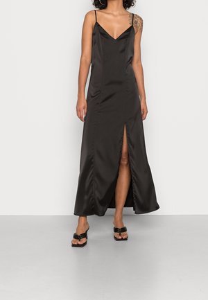 Kvinde iført en sort satin slipkjole med spaghetti-stropper og en frontslids ved låret, kombineret med sorte slidesandaler.