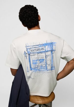 Göndör hajú férfi fehér pólóban, melyen egy "Vinyl Dreams" feliratú üzlet kék vázlata látható, egyik karján sötét zakót tart.