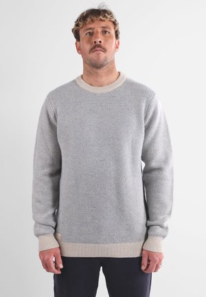 Homme avec des cheveux courts et bouclés et une moustache, portant un pull en laine texturé gris clair et un pantalon sombre, se tenant devant un fond blanc uni.