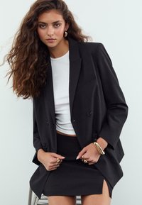 Frau mit langen lockigen Haaren, die ein schwarzes Blazer, einen schwarzen Rock mit Schlitz, ein weißes Oberteil sowie goldene Creolen, Ringe und ein Armband trägt und auf einem Hocker sitzt.