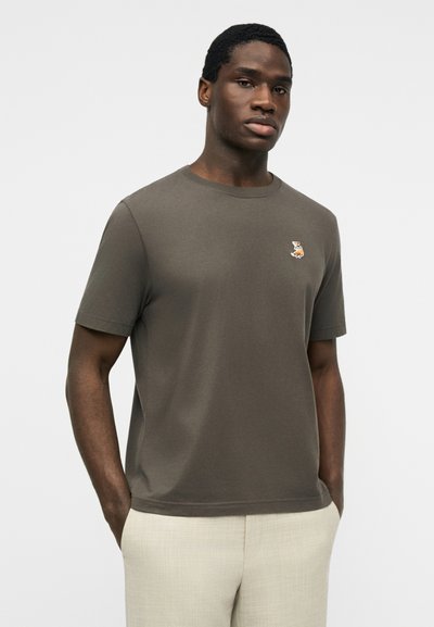 Άνδρας που φοράει κοντομάνικο χακί πράσινο μπλουζάκι με μικρό κεντητό λογότυπο σκύλου στο στήθος και ανοιχτό μπεζ παντελόνι.